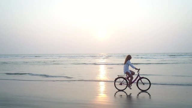 A young pretty girl riding on her bicycle at sunset. She rushes along the seashore beautifully reflected in the water on the sand. Flying camera moves near. Aerial view.