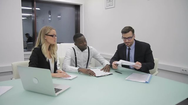 Male boss is explaining the market situation to african analytic and female manager. People are sitting at the table in office, with laptop on and businessman in suit and glasses is pointing the
