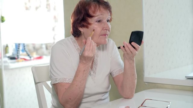 old woman makes a makeup in front of a mirror at home