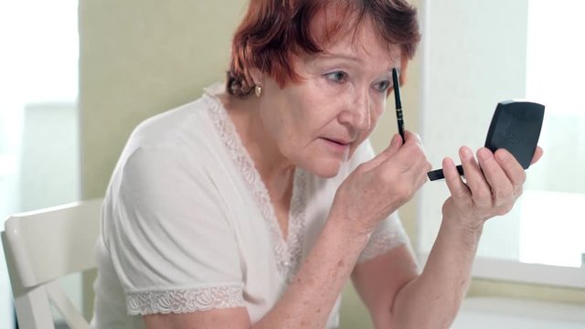 old woman makes a makeup in front of a mirror at home