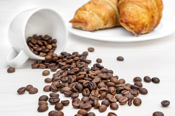 Coffee beans scattered from cup and two croissants on the table.