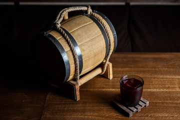 An underexposed horizontal image of a red alcoholic drink in a cocktail glass, on a wooden glass-holder on a table, a wooden barrel near by. Selective focus.