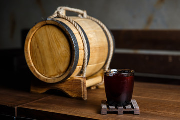An underexposed horizontal image of a red alcoholic drink in a cocktail glass, on a wooden glass-holder on a table, a wooden barrel near by. Selective focus.