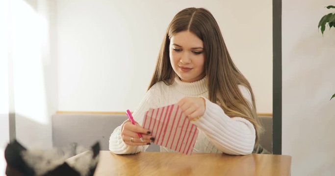 Brown-haired young women in white sweater write, smiling, get, read a Valentine's Day card and sign in reply in stylish cafe, home.