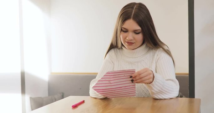 Brown-haired young women in white sweater write, smiling, get, read a Valentine's Day card and sign in reply in stylish cafe, home.