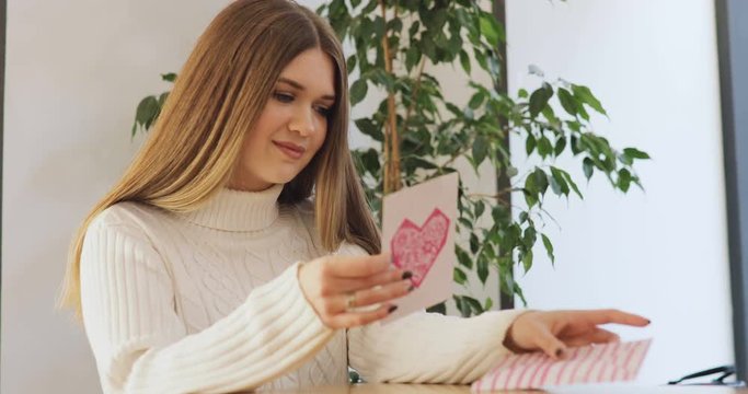 Brown-haired young women in white sweater write, smiling, get, read a Valentine's Day card and sign in reply in stylish cafe, home.