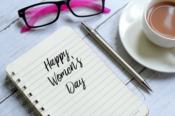 Top view of notebook written ' HAPPY WOMEN'S DAY' with a cup of coffee, pen and sunglasses on white wooden background.