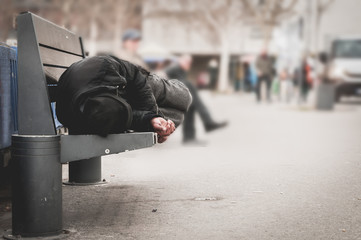 Poor homeless man or refugee sleeping on the wooden bench on the urban street in the city, social documentary concept, selective focus - obrazy, fototapety, plakaty