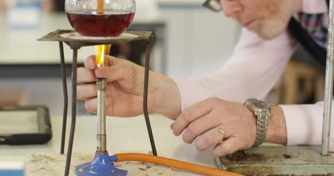 4K Teacher in school science class lighting Bunsen burner under a flask of liquid. Slow motion.