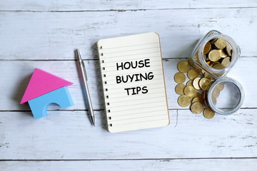 Top view of notebook written with 'HOUSE BUYING TIPS' with pen,house model and coins on white...