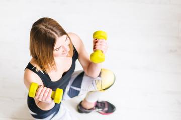 Top view of young woman  in black sportswear  holding dumbbells while sitting on the floor in bright fitness studio.