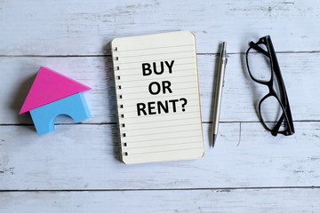 Top view of house model, sunglasses,pen and notebook written with 'BUY OR RENT?' on white wooden background.