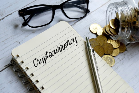 Sunglasses,jar of coins,pen and notebook written with 'CRYPTOCURRENCY' on white wooden background.
