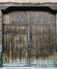 Old oak wood door with reinforcement nails, small door and iron knocker, with a wooden beam on top. It served for the passage of carriages to housing