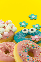 Many multicolored funny glazed donuts surrounded by artificial flowers on yellow background. Vivid hapyp contrast.