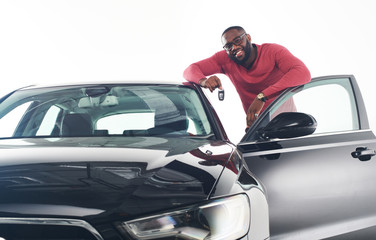 Happy handsome african man showing car keys in near his newly bought vehicle car smiling cheerfully copyspace owner ownership sales driving consumerism private taxi concept studio shot