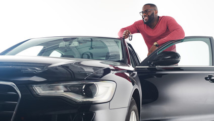 Feeling proud. Attractive young african man in stylish outfit standing near black luxury car model 2018 car and leaning on its roof.