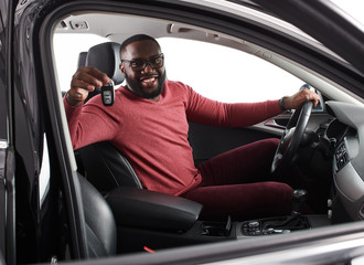 Happy handsome african man showing car keys in his newly bought auto smiling cheerfully sitting in...