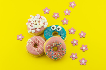 Many multicolored funny glazed donuts surrounded by artificial flowers on yellow background. Vivid hapyp contrast.