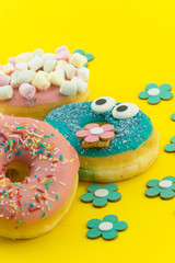 Many multicolored funny glazed donuts surrounded by artificial flowers on yellow background. Vivid hapyp contrast.