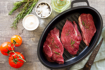 Fresh meat. Raw steak beef on a cast-iron grill frying pan, tomatoes, olive oil, spices and fresh rosemary on the kitchen table. Top view.