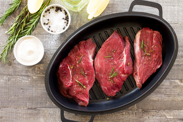 Fresh meat. Raw steak beef on a cast-iron grill frying pan, olive oil, spices and fresh rosemary on the kitchen table. Flat lay. Top view.