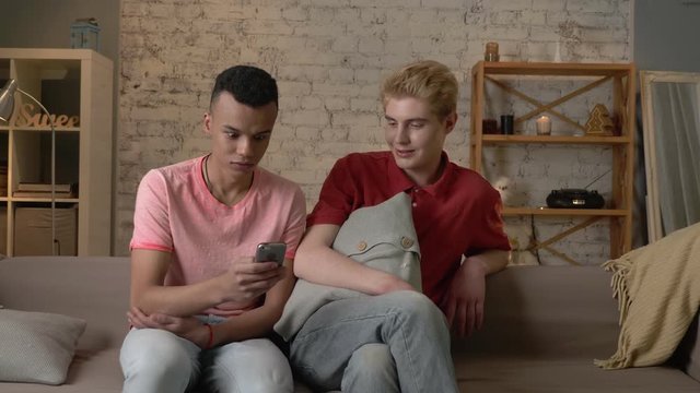 Two international friends of a homosexual are sitting on the couch and watching shocking, mercenary pictures on the smartphone. Home cosiness, family, internet concept. 60 fps