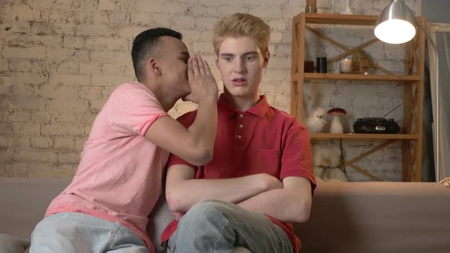 Two young homosexual boys are sitting on the couch, african guy with short hair tells the secrete to his partner, the blonde is shocked 60 fps