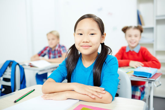 Little diligent girl sitting by her desk with two classmates on background