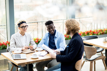 Young intercultural colleagues listening to report of their co-worker while sitting by table in cafe