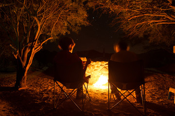 Couple sitting at burning camp fire in the night. Camping in the forest under starry sky, Namibia, Africa. Summer adventures and exploration in the african National Parks. Selective focus on fire.