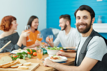 Happy bearded man with toothy smile sitting by festive table with his friends