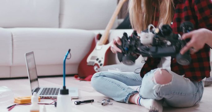 Young attractive teen girl listening to music and dancing while assembling toy RC car at home, modern interior. 4K UHD