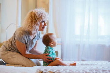 Blond curly female nanny with infant baby boy sitting on the bed in cozy interior bedroom. Side view. Real people life and different generations concept.