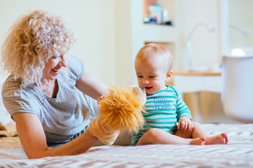 Blond curly female nanny playing with happy infant baby boy while sitting on the bed in cozy interior bedroom. Side view. Real people life and different generations concept.