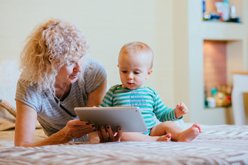 Little cute infant baby boy with curly blond female nanny working with tablet while siting in bedroom at cozy home interior. Real people life and different generations concept.