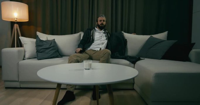 Young adult bearded Caucasian male sits on couch, turning on TV screen. 4K UHD