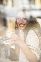 Cheerful young casual lady drinking her coffee in a coffee shop, enjoying some free time for herself, through the window shot