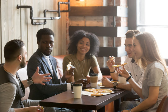 Multi-ethnic african and caucasian friends talking eating pizza in pizzeria, diverse young people enjoy italian food at meeting, multiracial students having conversation during lunch together in cafe