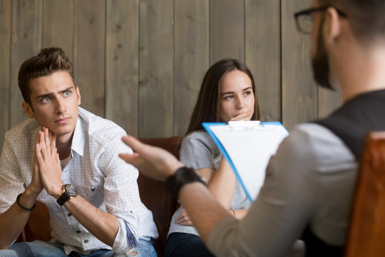 Frustrated Young Man Listening To Psychologist While His Offended Girlfriend Sitting Apart, Family Counselor Talking To Unhappy Couple Solving Problems In Relationships At Counseling Therapy Session