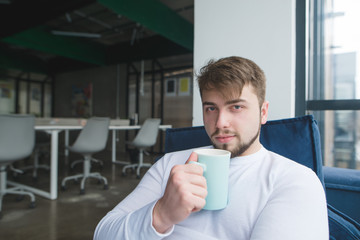 Portrait of a young man with a cup of drink in his hands at the background of the office. A man with a cup of coffee resting in the office on the couch.