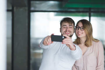 Smiling man and girl make selfie indoors. Beautiful pair of office workers to make selfie smartphone. Happy employees are photographed on a smartphone in the office.