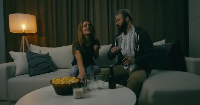 Young adult Caucasian couple sit on couch at home, watching TV, eating popcorn, drinking beer. 4K UHD