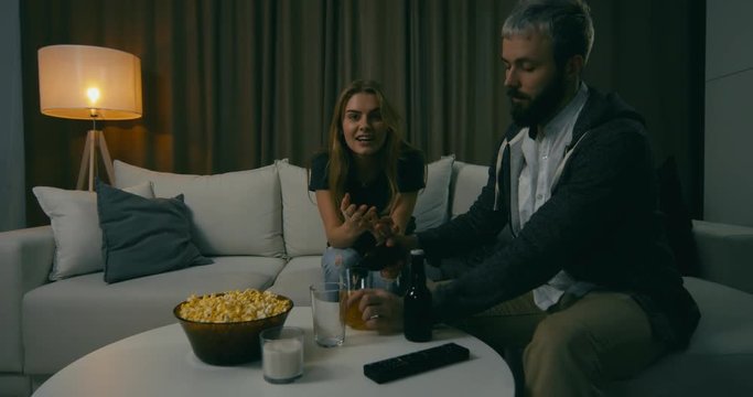 Young adult Caucasian couple sit on couch at home, watching TV, eating popcorn, drinking beer. 4K UHD