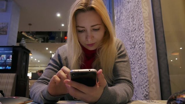 4k Pretty blond hair young girl looks photos at mobile phone in cafe