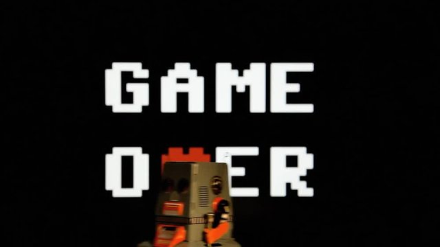 A tin toy robot moving in front of a Game Over screen (blocky C64 8-bit characters, a heart replacing the letter V).
