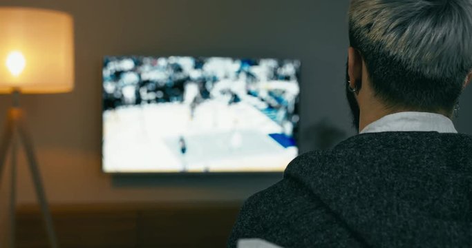 RL DOLLY Back view of Caucasian male sitting on sofa, watching sport basketball game on a TV screen. 4K UHD