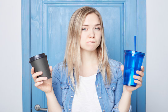 .Doubtful hesitant intriguing blond female curves lips, has thoughtful expression, can`t decide or make choice between addiction and health, dressed in blue casual shirt. Facial expressions concept.