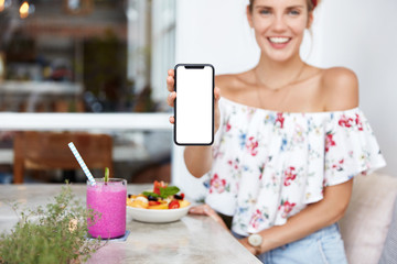 Cheerful restful woman sits at restaurant, enjoys fresh fruit and beverage, holds smart phone with blank white screen for your promotional text or advertisment. People, technology, lifestyle concept