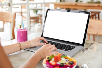 Successful female freelnacer uses laptop with blank screen during vacations in exotic country, does remote job, surrounded with fresh smoothie and fruit salad, enjoys free internet connection in cafe.